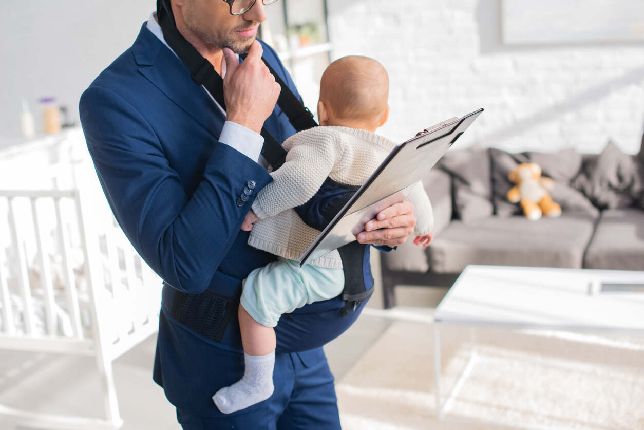 A focused father in a suit holds his baby in a carrier while reviewing documents on a clipboard, symbolizing the importance of establishing legal paternity for fathers and children in Iowa.