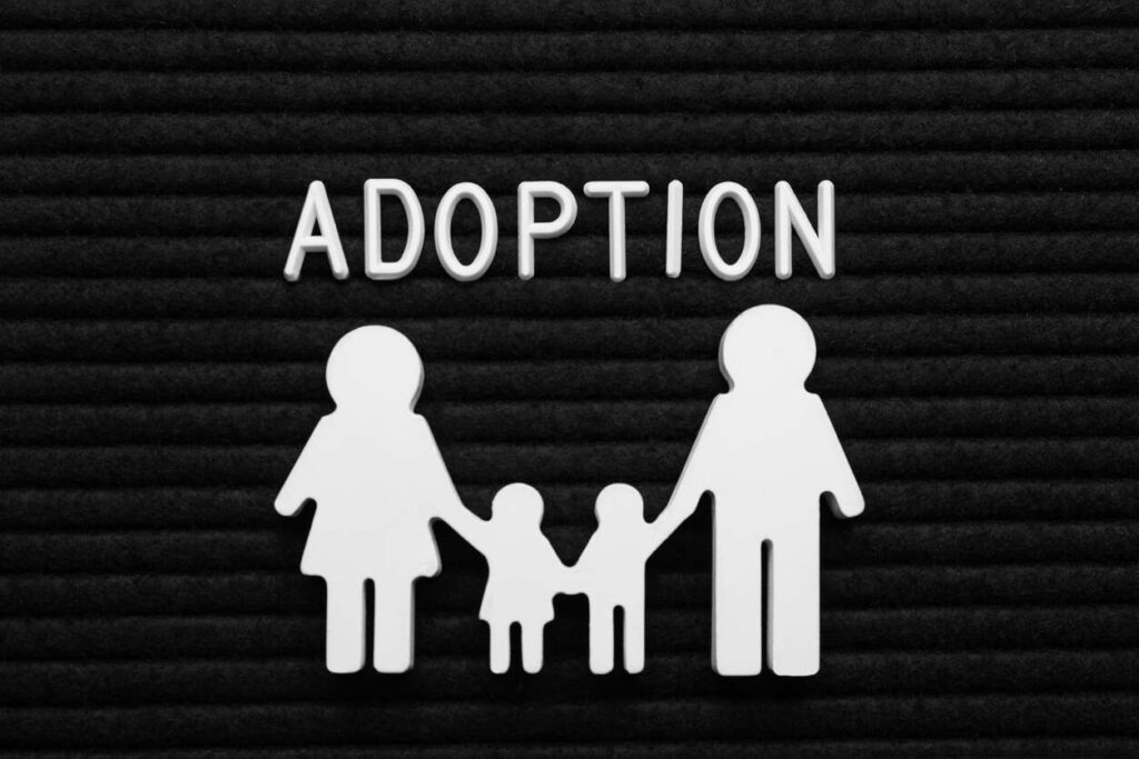 White cutout silhouettes of a family holding hands positioned below the word 'ADOPTION' on a black board, symbolizing the legal process and requirements for stepparent adoption in Des Moines.